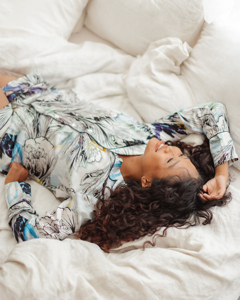 Image is of Indonesian model Dina laying face up on plush white bedding and laughing. She is wearing the long sleeved Nilofar print Iris silk Pajama set.