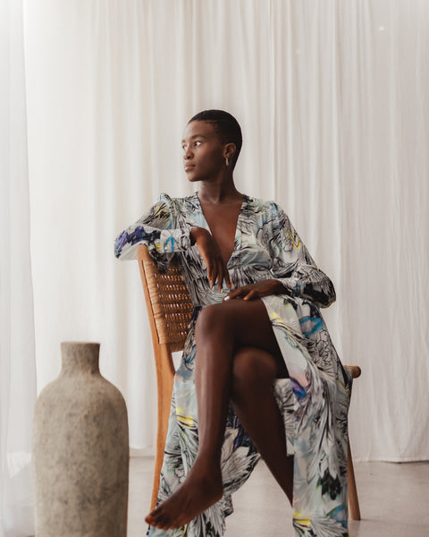 Image is of South African model Thoko. She is sitting elegantly on a wooden chair with one legged crossed over the other. She is wearing the Coco Silk Robe in Nilofar print, which stands out beautifully against her dark brown skin.