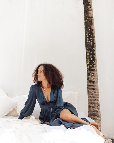 Image is of a female model wearing a lustrous navy blue silk robe on a plush white bed. 