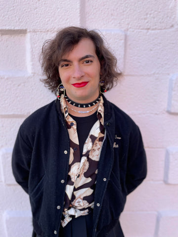 a white trans femme person with medium long hair, wearing lipstick, a choker necklace, a tshirt, an unbottoned collared shirt with a print, and a black cardigan, standing against a white brick wall (artemis)