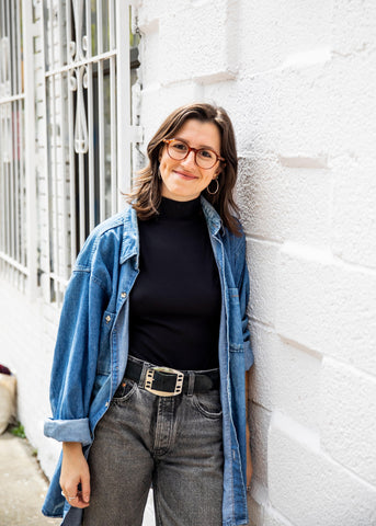 Carolyn, a white female presenting queer person with shoulder length dark hair wearing glasses, gray jeans, a belt, a black turtleneck, and a chambray open button-down shirt, leaning against the white brick wall of Shag in Brooklyn