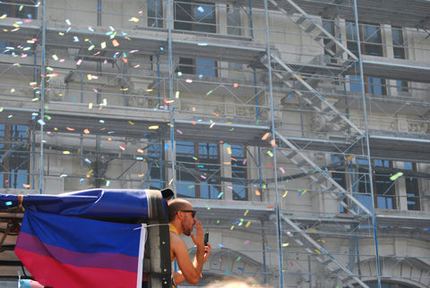 bi flag on a float at a pride parade, with a person sticking out of cannon type structure, paper confetti in the air, and a city building in the background