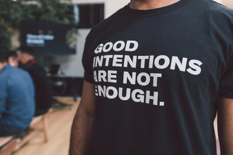 black masculine presenting torse (head cut off from image) wearing a black tshirt with words "good intentions are not enough." printed in white, standing at an angle in a brightly lit space with hardwood floors