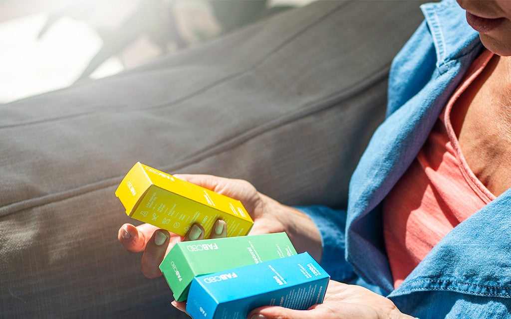A woman holds three boxes of different flavored CBD as she sits on a sun-drenched couch