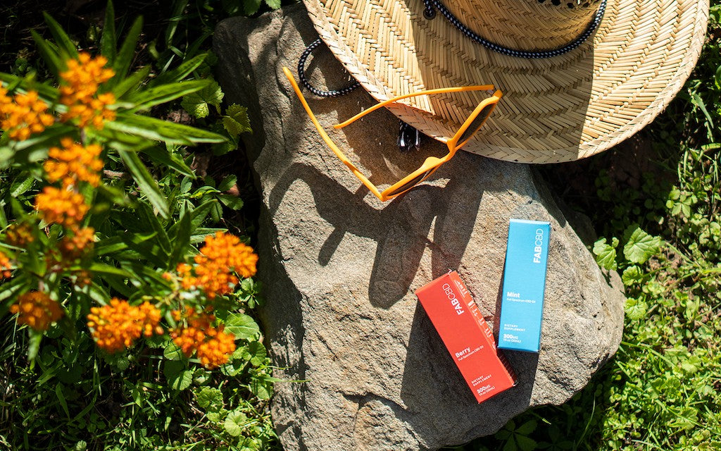 A straw hat, a pair of sunglasses, and two boxes of CBD oil rest on a sunny rock surrounded by grass and orange flowers.