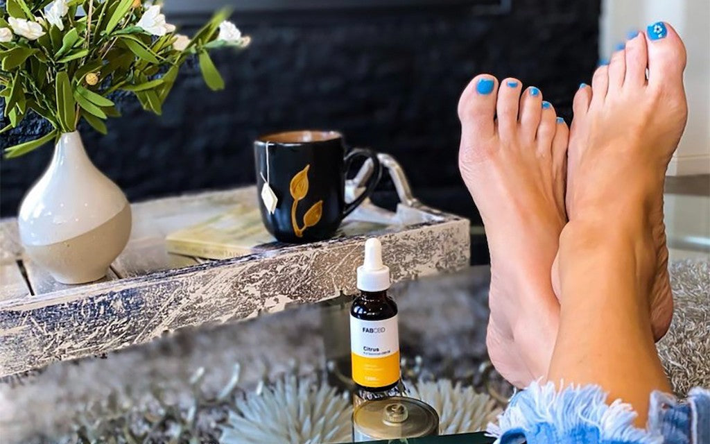 A woman's bare feet are propped up on a stone table next to a bottle of CBD oil