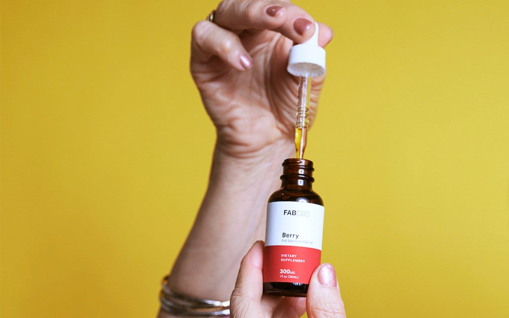 hands hold a bottle of CBD and lift the dropper part-way out of the bottle in front of a solid yellow backdrop.