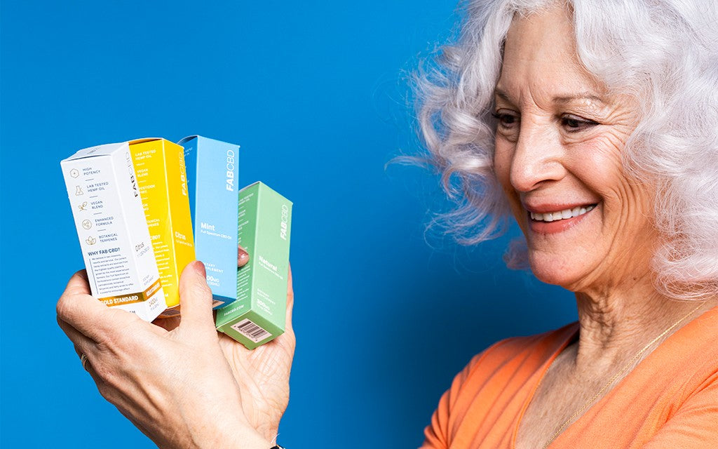 A woman with wavy white hair smiles as she displays multiple colorful boxes of FAB CBD oil in front of a blue backdrop