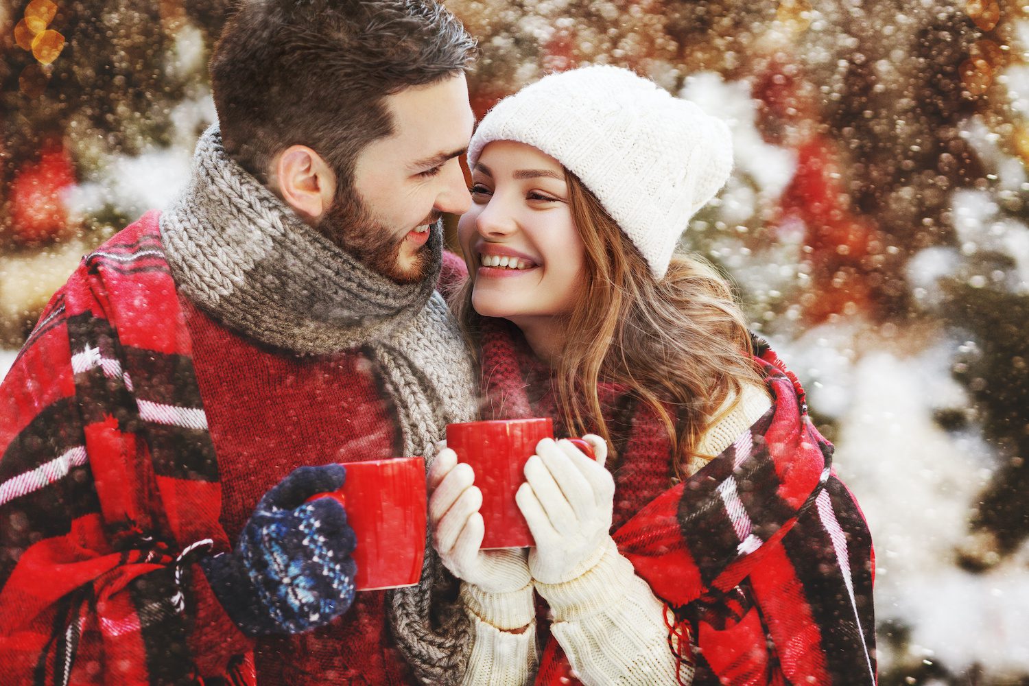 A couple cozies up toether wrapped in warm winter gear and holding steaming mugs of cocoa