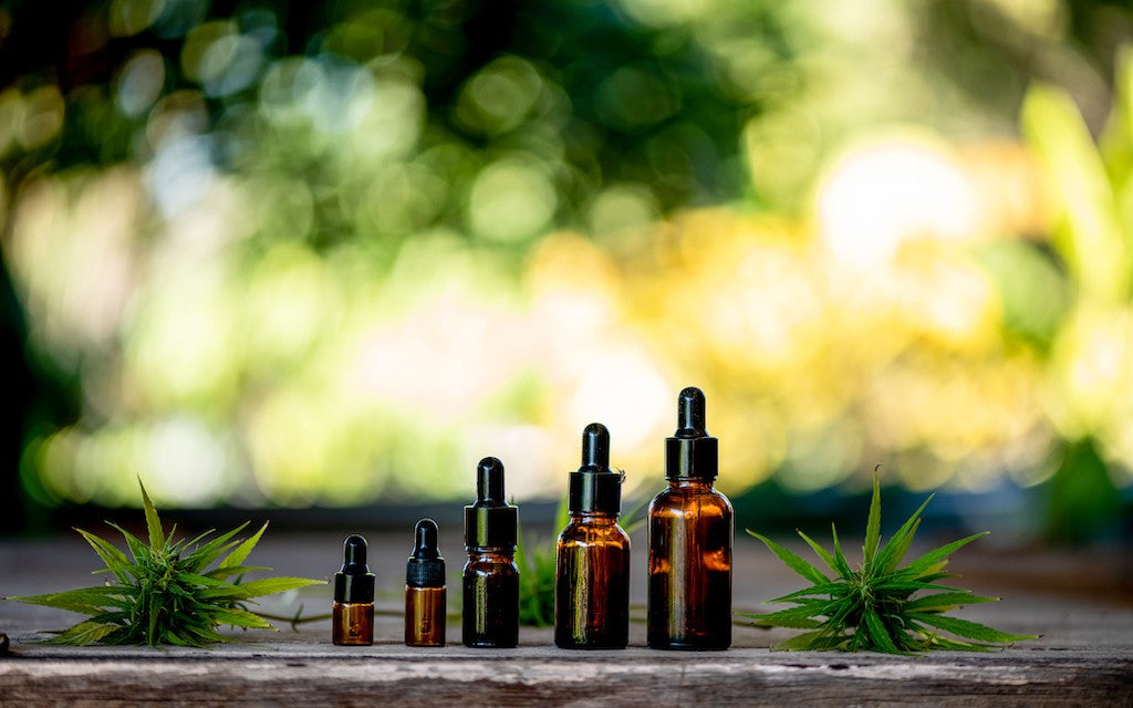 A row of successively larger bottles of CBD lined up on a wooden surface outside with hemp leaves laid down next to the bottles