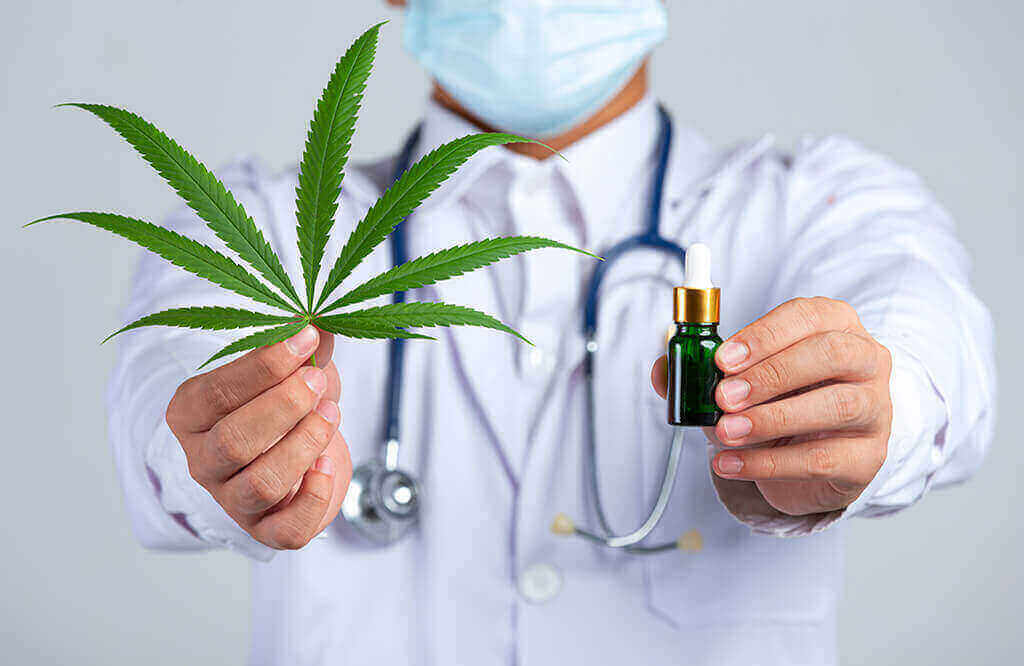 A man in a doctor's coat and wearing a stethoscope holds up a hemp leaf and a tincture bottle