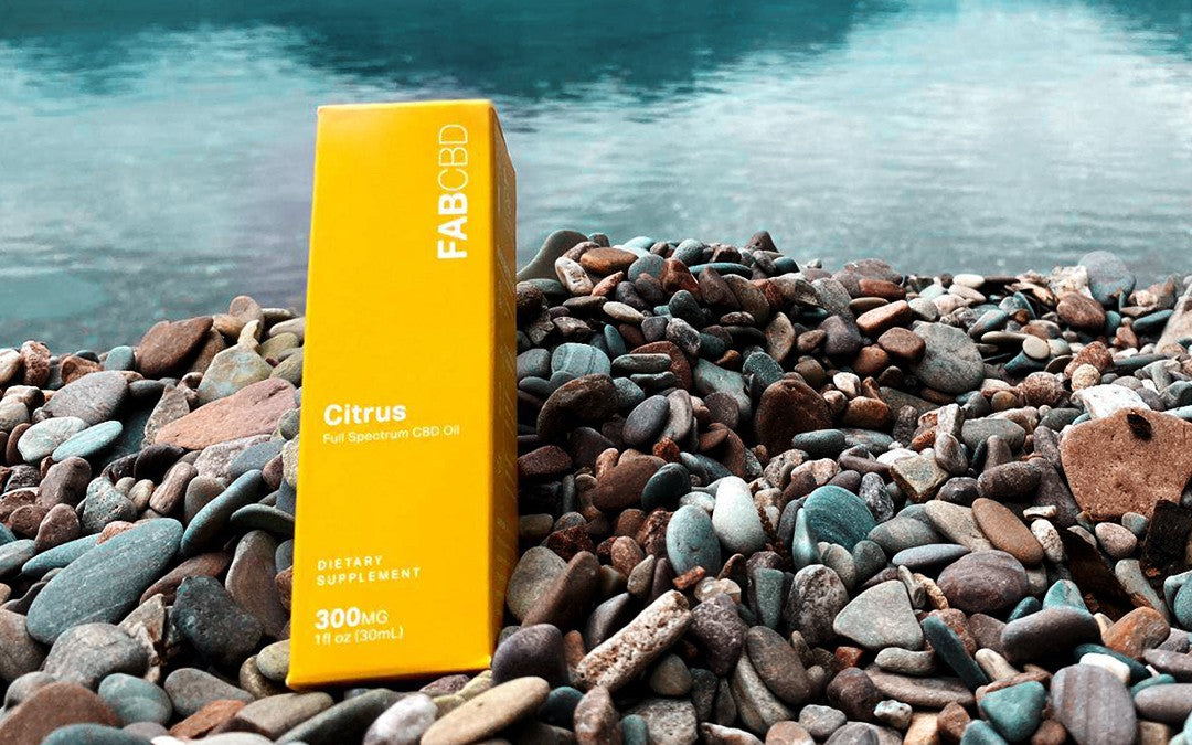 A yellow box of FAB CBD citrus oil resting on colorful pebbles along a river bank.