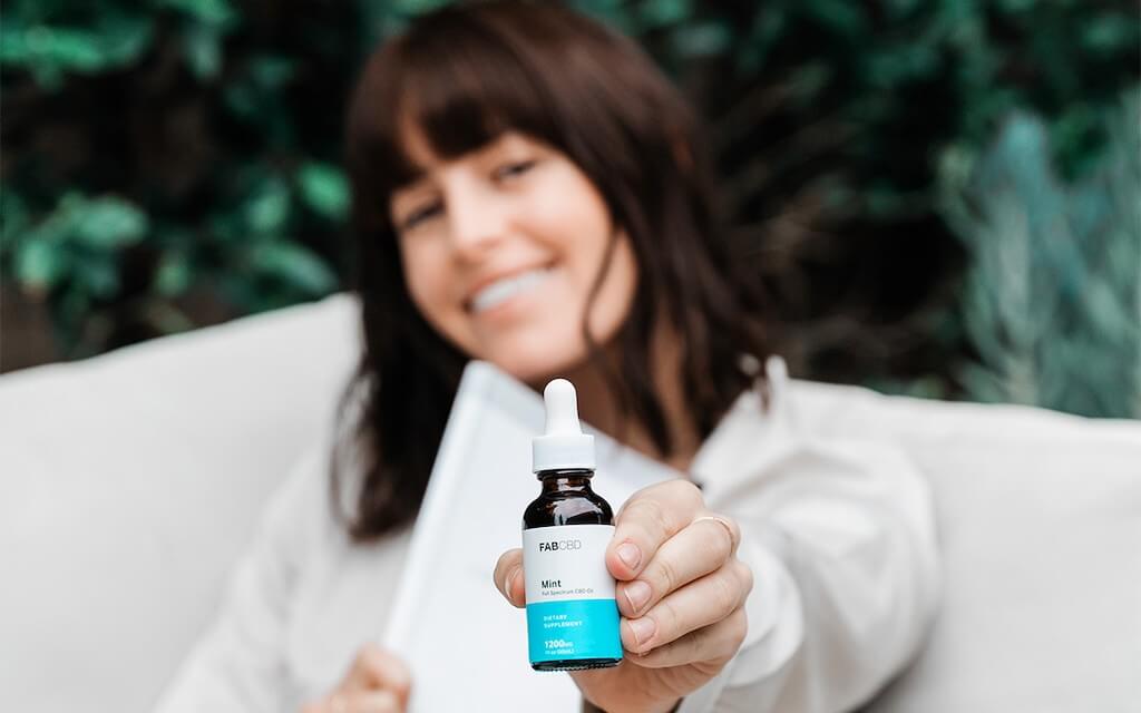 A smiling woman holds a book to her chest and a bottle of CBD up to the camera