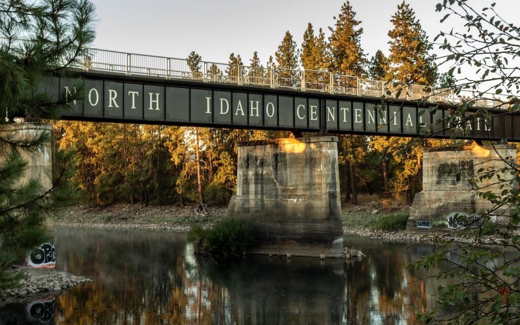 A picturesque bridge over a tree-lined river says "North Idaho Centennial Trail"