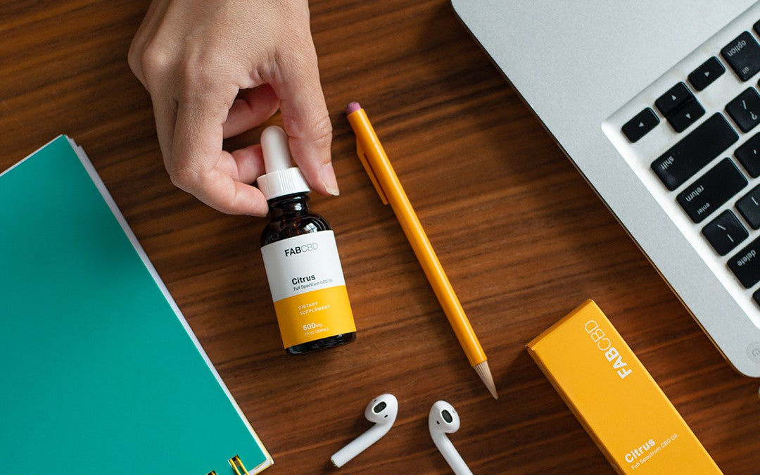 A hand reaching for a bottle of CBD oil next to a laptop, some headphones, and a pencil. 