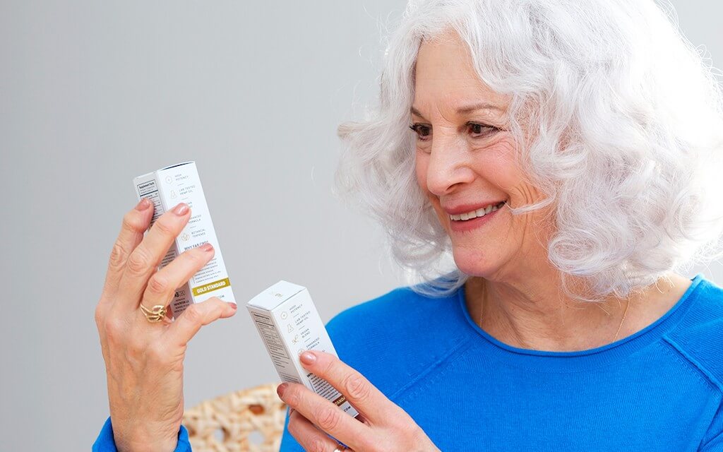 A woman with chin-length white hair and wearing a blue shirt holds up to boxes of CBD oil and reads the ingredients with a smile