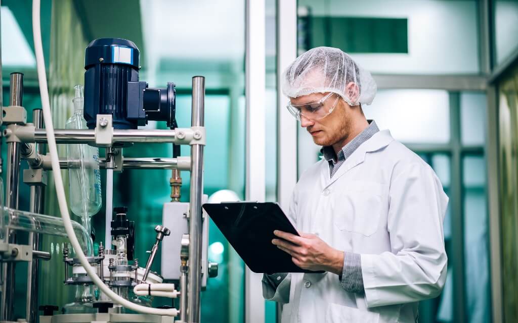 A scientist in a lab coat writes on a clipboard while standing next to equipment in the lab
