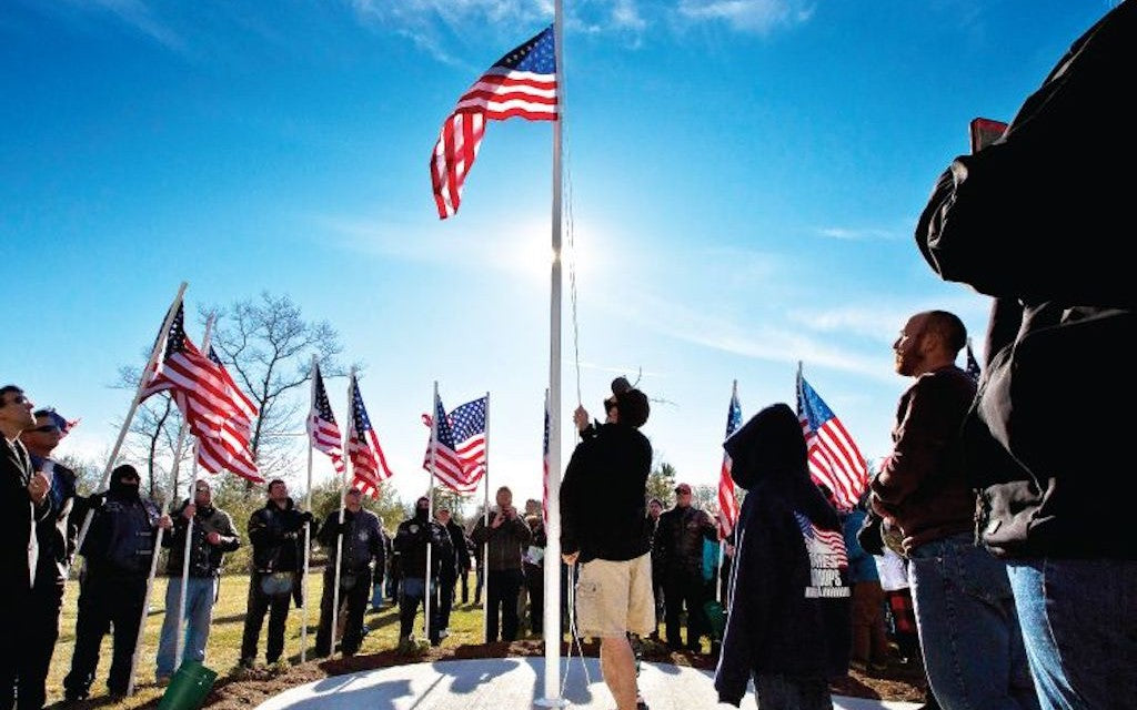 People standing near a number of flag poles with American flags waiving in the wind and sun.