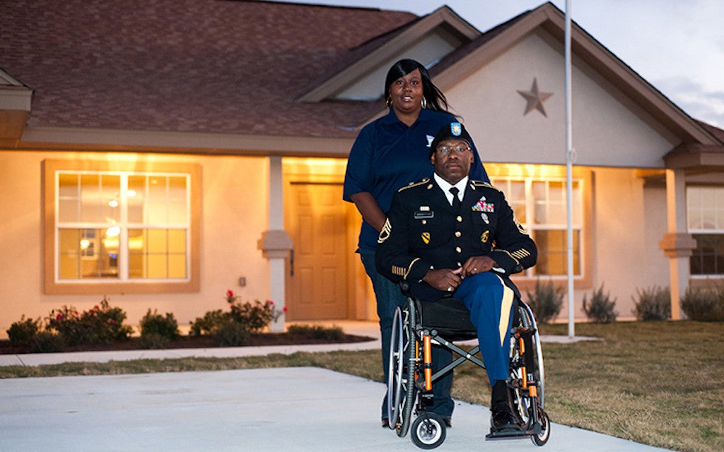 A woman stands behind a Veteran in a wheelchair on the driveway of a home built by Homes For Our Troops