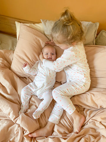 Egy kislány és egy kisbaba fekszik az ágyon egyforma szíves bambusz kezeslábasban