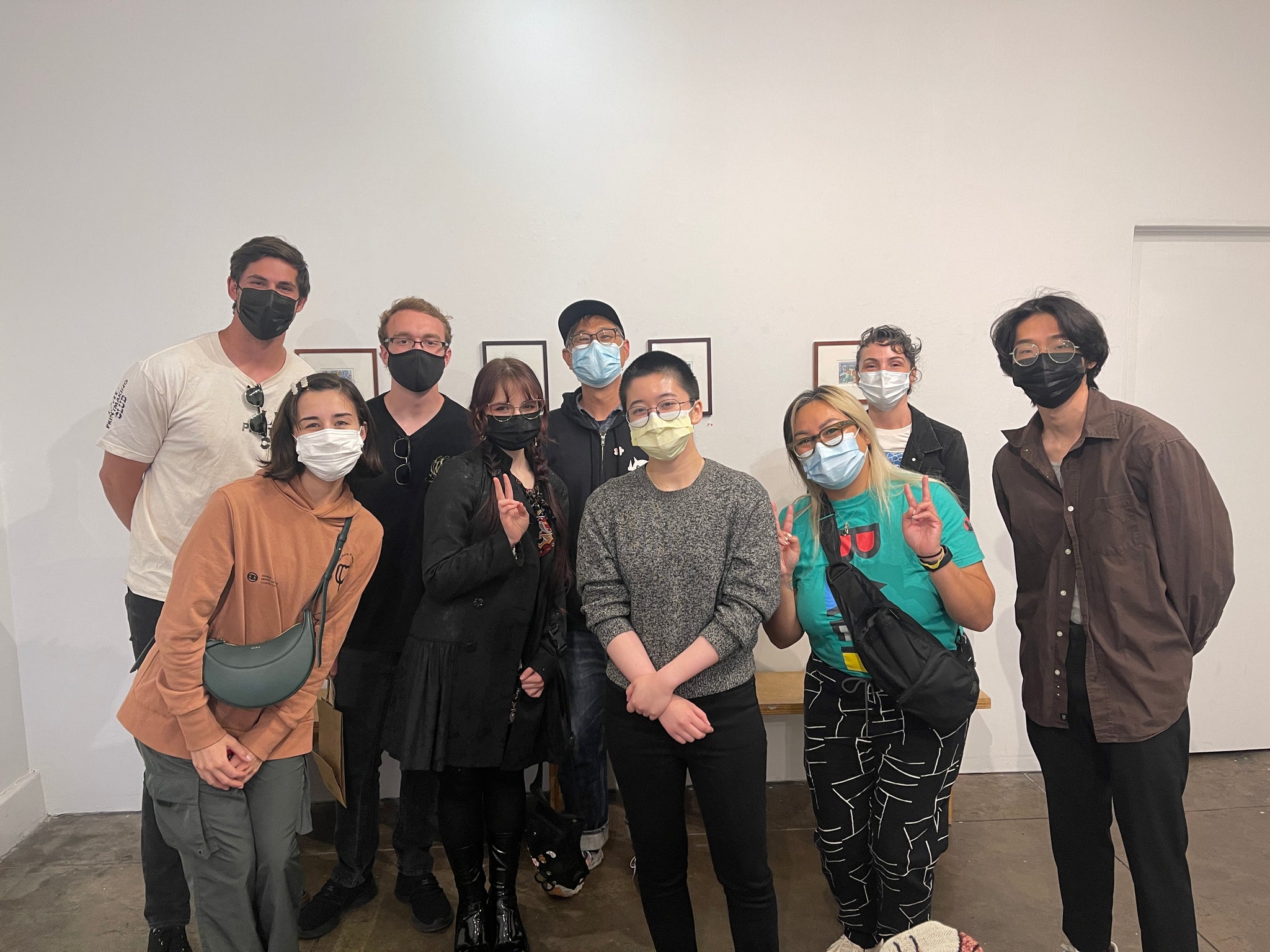 Group image of nine people in a mostly blank wall gallery setting. There are tiny pieces of art on the wall.