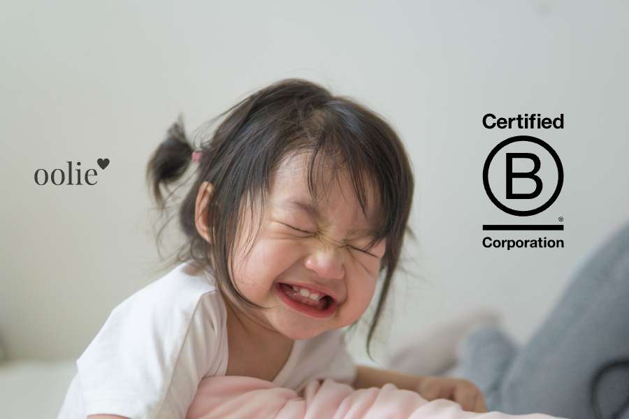 Photography of a baby girl grinning and laughing, with the Oolie logo and a symbol representing Certified B Corporation