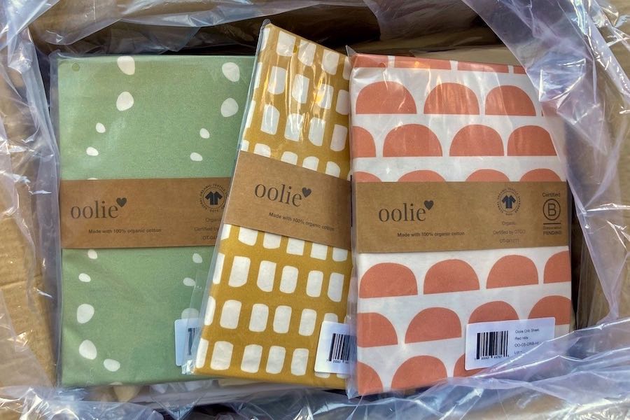 Three Oolie organic cotton crib sheets, with graphic prints, shown fully packaged in an open box.