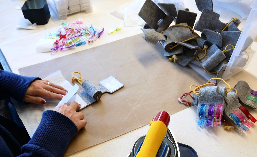 A person's hands, reaching over a work surface, assembling an Oolie Shoosh door silencer and safety bumper, with materials and tools nearby.