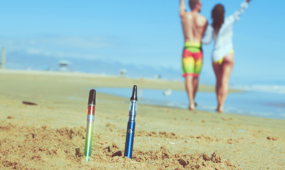 Man and woman walking away from Exxus vapes buried in the sand at the beach with natural lighting