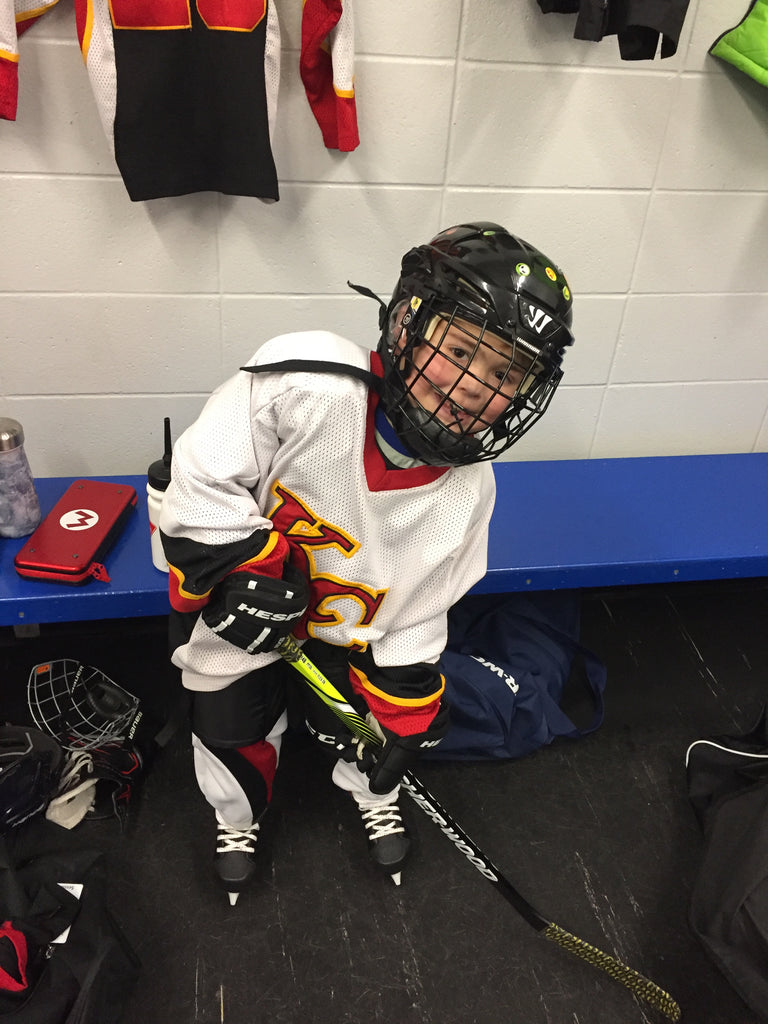 Photo of Nate in his hockey equipment in the locker room, he has his stick down and looks ready to play. 