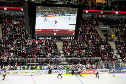 An LED Digital Display in a hockey arena, showing a close up shot of the hockey game that's proceeding below it. The stadium seats are full of fans.