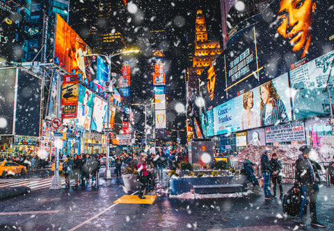 Picture of Times Square at night while it snows. LED Digital Displays are playing different ads while people walk below.