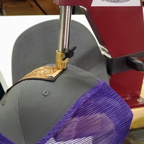A wood and metal plate patch hat being riveted. This trucker hat is charcoal and purple. The decoration is etched alder wood veneer laminated to a metal plate and riveted to a hat.