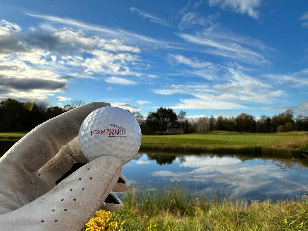 Hand holding J. Campoli & Sons, Inc. branded golf ball in front of a pond