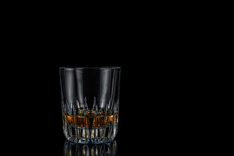 A rocks glass with about of finger's worth of whiskey in it sits in front of a black background.