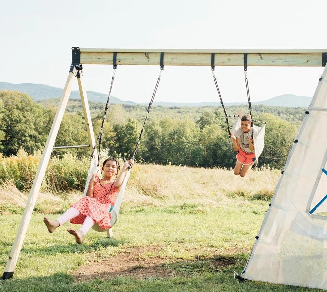 Two children swinging joyfully on swings in a lush, scenic outdoor setting.