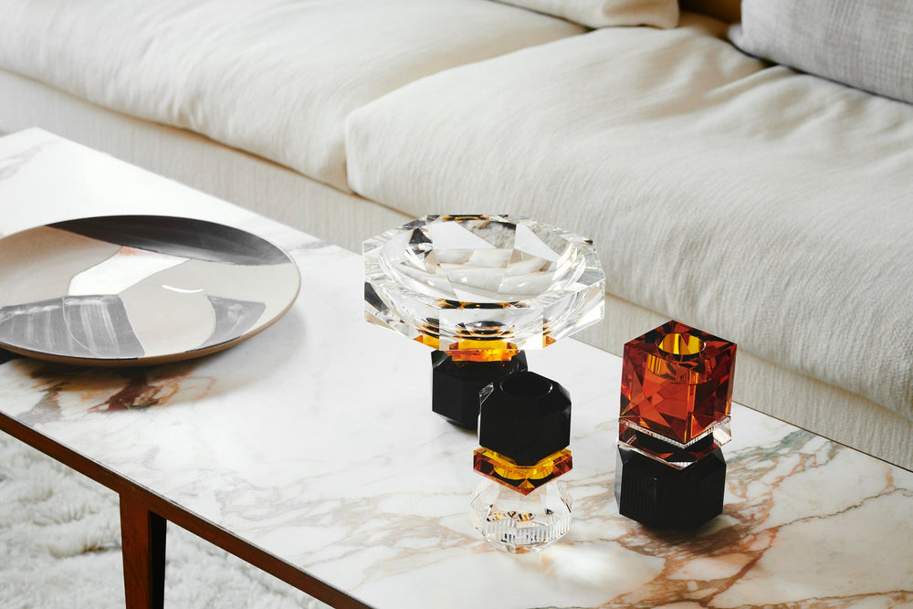 Arrangemang med ljusstakar och en skål i färgad kristall, på ett soffbord i marmor med en soffa i bakgrunden.