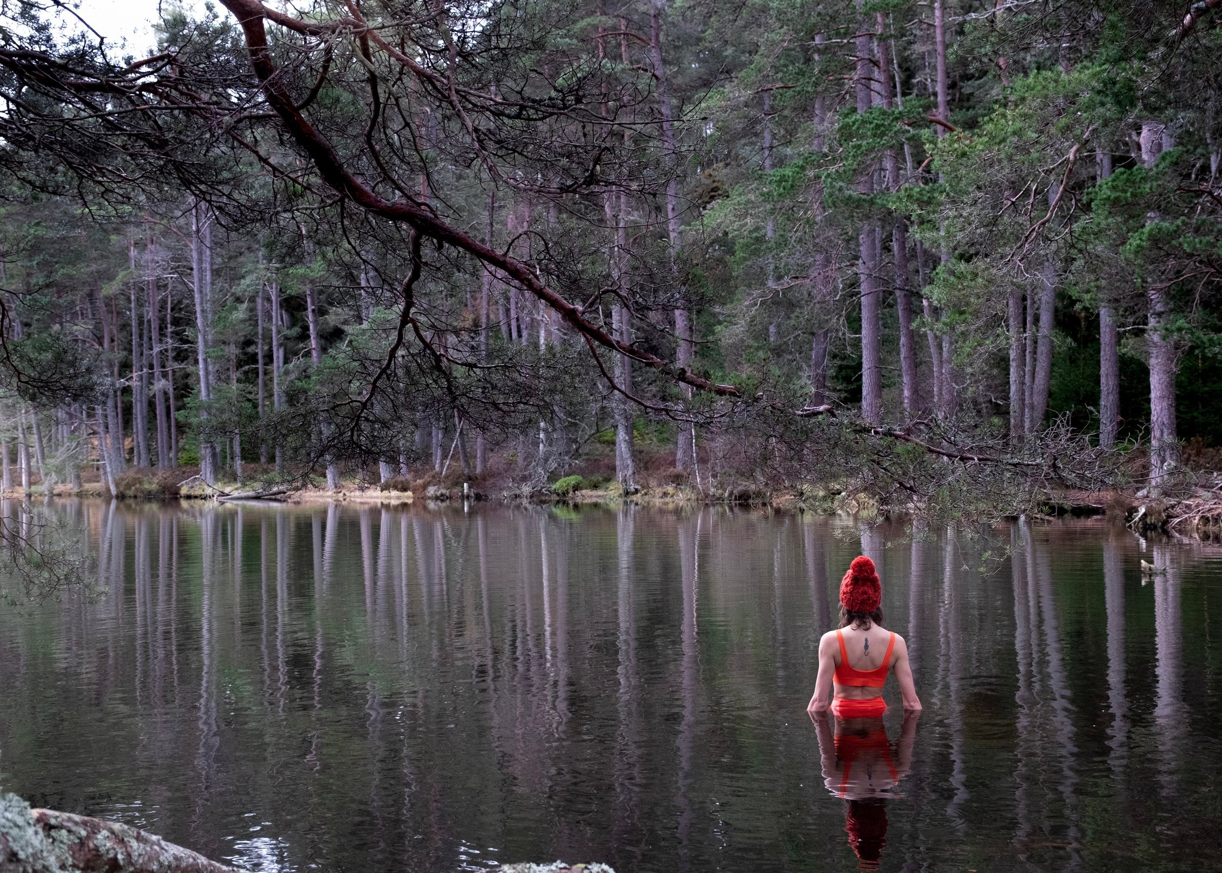 A woman standing waist deep in a loch surrounded by forest. She is facing away wearing an orange bikini and red bobble hat.