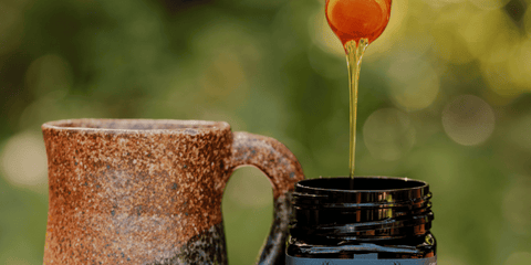 manuka honey dripping from honey spoon into gather by manuka honey jar with a tea cup next to it