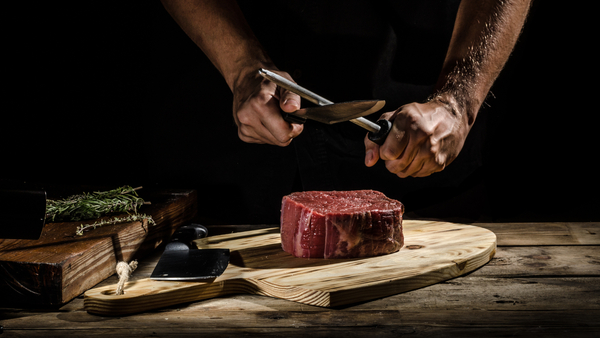 A person sharpens their knife over a cutting board with a large piece of protein ready to be trimmed and cooked for a bodybuilder's meal.