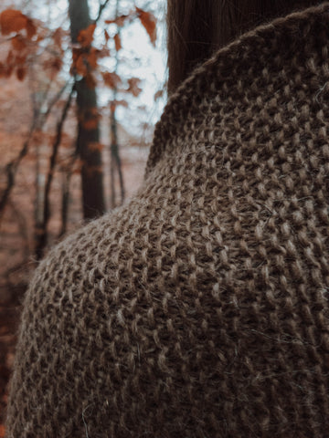 A close-up of the Tåkesti Shawl, knit in a medium brown Nutiden unspun yarn colorway, highlighting a textured stitch pattern with slipped and knit stitches. In the background, vibrant fall colors and trees add to the scenic view.
