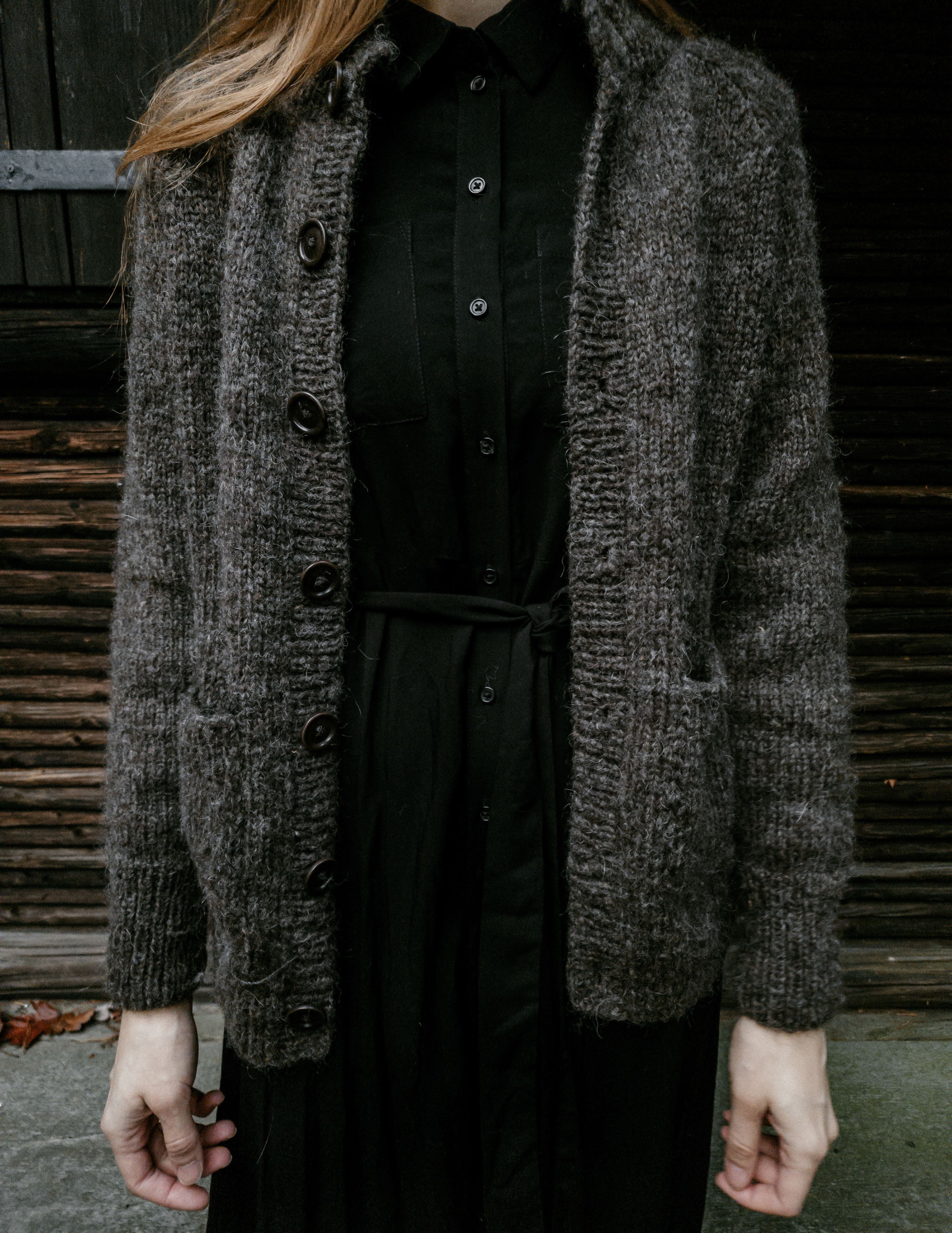 Image of knitwear designer Lív Ulven wearing her handknit rustic dark grey Lo·ki Cardigan, crafted with unspun Nutiden yarn. The background features an old, dark brown wooden cabin wall.