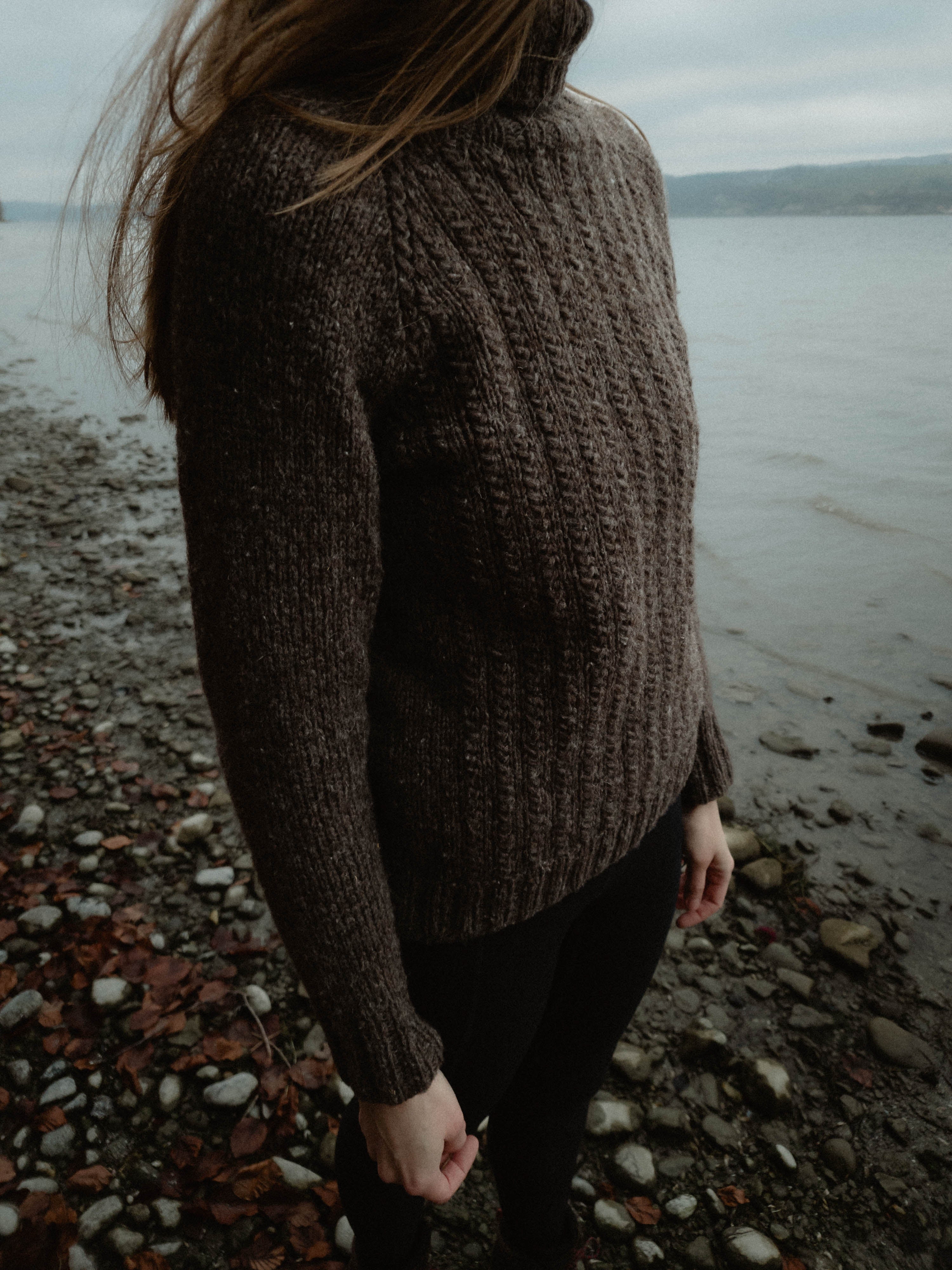 Image of knitwear designer Lív Ulven wearing a natural dark grey Fisherman's Raglan sweater with textured front, handknit in Manchelopi unspun yarn, standing on a stony beach. The backdrop showcases a foggy large body of water.