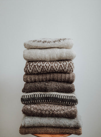 A stack of handknit sweaters, designed by Lív Ulven and crafted from unspun yarn, arranged on a small round table against a white wall. The sweaters feature intricate textures and captivating colorwork designs.
