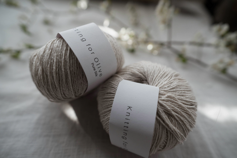 Two balls of Knitting for Olive - Pure Silk in a natural beige colour displayed on a linen cloth on a table.