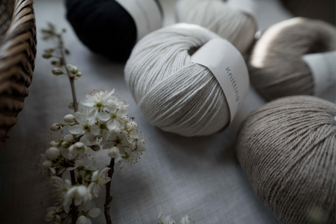 Balls of Knitting For Olive - Cotton Merino yarn displayed on a natural background. There is a branch with white flowers on the table near the yarn and an edge of a woven basket is visible. The balls of yarn are in various colours of beige, white and green.