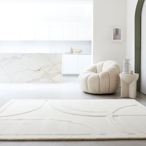 A minimaluxe room with elements of luxury and minimalism. This room is simple and calming. There is a simple kitchen i the background with a cool toned marble effect. The furniture and decor are soft with round curved edges. There is a mix of texture in this room. The large cream rug is simple and monochromatic but features a textured 3D design with curves.