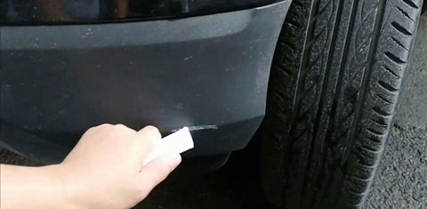 A person using a heat gun to carefully apply heat to the scratched area of a black plastic bumper, causing the plastic to soften and the scratches to disappear.
