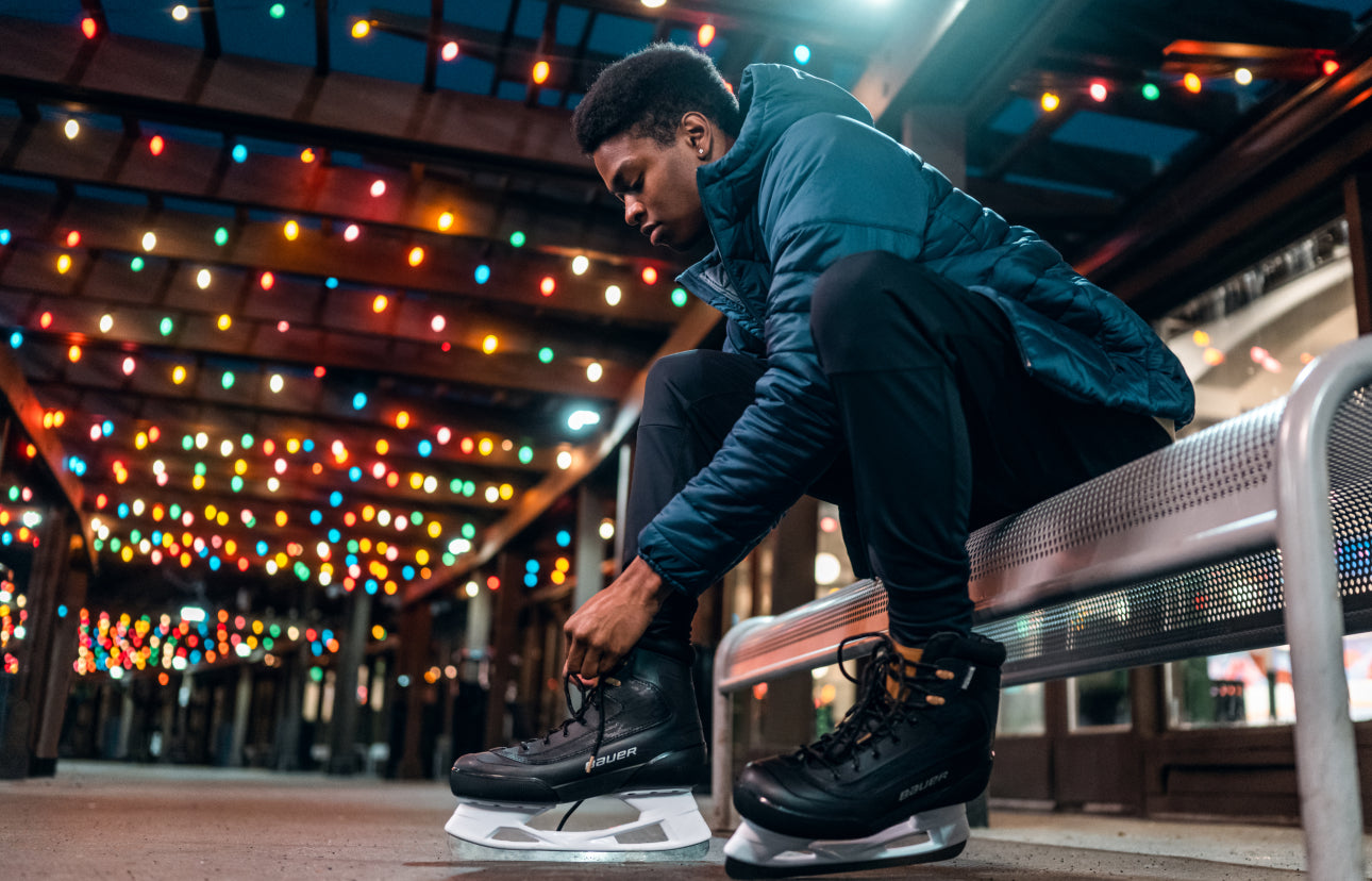 man sitting on an outdoor bench lacing up a pair of skates under colorful string lights