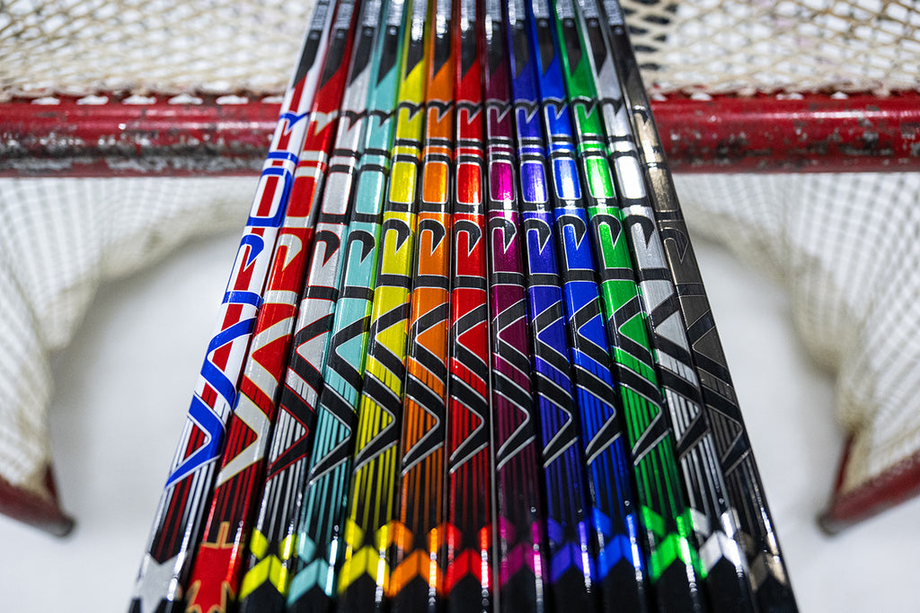 close up of a dozen colorful custom hockey sticks lined up on top of a hockey net